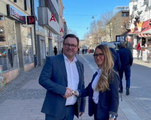 Patrick Hansson, en mann med briller i blå dress og hvit skjorte til venstre håndhilser på Rebecca Moberg, en kvinne med langt blondt hår, biller og blå dressjakke og hvit skjorte ute på en gågate i Sverige.